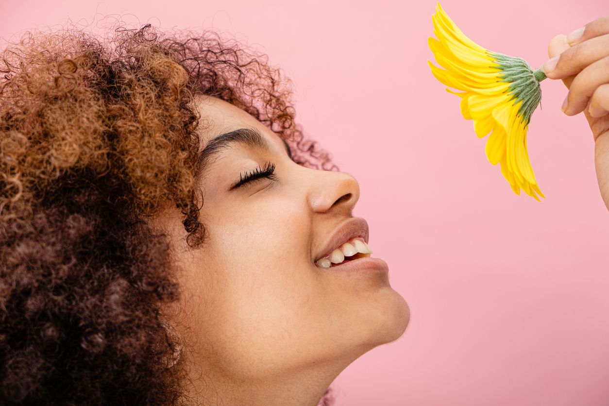 A young woman with curly hair and radiant skin holds up a fresh flower to her smiling face